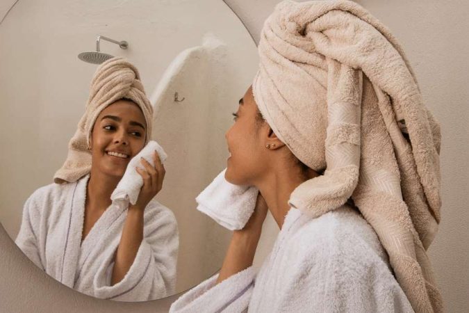 Une jeune femme en peignoir et serviette sur la tête se regarde dans un miroir rond et essuie délicatement son visage avec une serviette blanche.